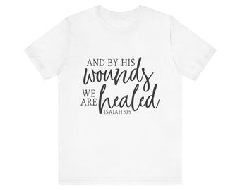 May include: White t-shirt featuring the text "AND BY HIS wounds WE ARE healed" in black script. The text also includes "ISAIAH 53:5". This is a casual wear item.