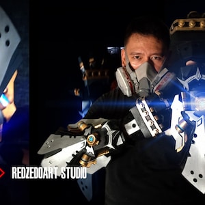 以下が含まれることがあります： 黒いシャツを着て、ガスマスクをつけた人が、青色の光がついた、精巧な銀と黒の機械的な腕を持っています。腕には、大きな銀色の刃と、小さな金色の刃が付いています。背景は濃い青色で、明るい青色の光が当たっています。