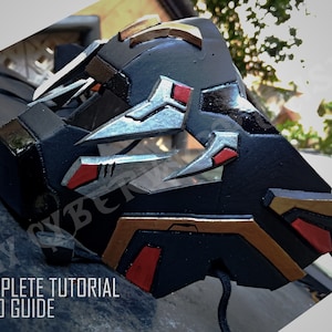 May include: A black, silver, and gold mask with red accents. The mask has a geometric design and is made of a hard material. The mask is attached to a black string.