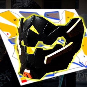 Könnte beinhalten: Eine schwarz-gelbe 3D-gedruckte Maske mit geometrischem Design. Die Maske wird vor einem weißen Papier mit einem Muster für die Maske gehalten.