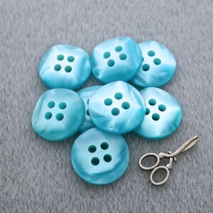May include: A collection of eight round, light blue buttons with a glossy finish, each featuring four central holes arranged in a square pattern. Beside the buttons lies a small, metallic, antique-style pair of scissors with a detailed handle.