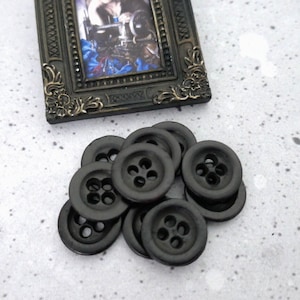May include: A collection of dark gray, four-hole buttons. The buttons are round and have a matte finish. In the background, a small, ornate picture frame with a vintage image of a person sewing.
