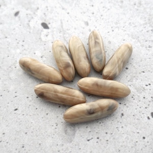 May include: A collection of seven light brown, oval-shaped beads with a marbled appearance. The beads are arranged on a light gray, speckled surface. The beads have a smooth, polished texture and varying shades of brown.