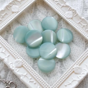 May include: A collection of pale blue, round buttons with a pearlescent sheen, arranged inside a white, ornate picture frame. A pair of small, silver scissors rests near the frame. The background is a white, textured knit fabric.