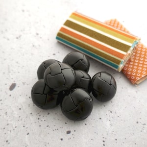 May include: A group of eight black leather buttons with a textured surface. The buttons are arranged in a pile.