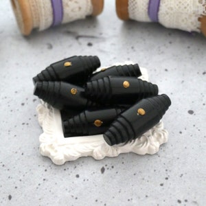 May include: A group of black, wooden, button-like beads with gold accents. The beads are arranged in a pile on a white, decorative, framed surface.