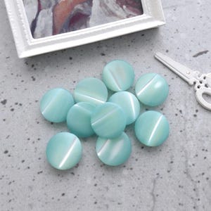 May include: A collection of ten round, smooth, light turquoise buttons with a subtle pearlescent sheen. Beside the buttons lies a small, white, decorative metal scissor-shaped object, and above them is a framed painting of a person in white clothing.