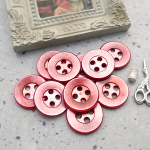 May include: A collection of red buttons with five holes each, a small spool of thread, and a pair of silver scissors. A small framed picture is in the background. The buttons are arranged on a speckled gray surface.