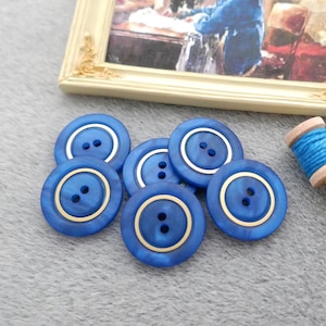 May include: A collection of blue buttons with gold rims and two holes each. A small framed picture and a spool of blue thread with gold scissors are also present. The buttons are a sewing or craft supply.
