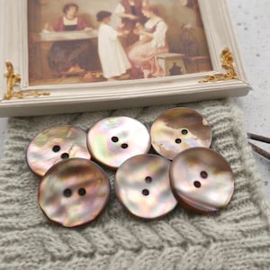May include: A collection of iridescent, round buttons with two holes each, arranged on a gray knitted fabric. A framed painting with gold accents rests above the buttons. Two small, dark brown knitting needles are also visible.