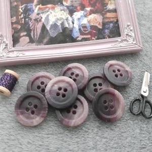 May include: A collection of purple buttons with four holes, a spool of purple thread, and small black scissors. A framed picture of two women sewing is in the background. The buttons are various shades of purple and gray.