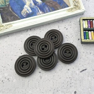 May include: A collection of dark brown, circular buttons with concentric ring detailing. Each button has two holes for sewing. A small framed painting and a miniature sewing kit are in the background.
