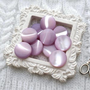 May include: A collection of 10 lavender-colored buttons with a pearlescent finish, displayed in a white ornate frame.