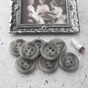 May include: A collection of gray and white marbled buttons with four holes, alongside a small framed antique print depicting three women. A small spool of thread is also present, suggesting a sewing or craft theme.