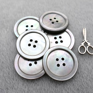 May include: A collection of iridescent, round buttons with four holes each, and a small pair of silver scissors. The buttons have a pearlescent sheen and are arranged on a gray surface.