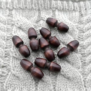 May include: A collection of dark brown wooden toggle buttons. Each button has a rounded, elongated shape with a narrow center. The buttons are scattered on a light gray, textured fabric, likely a knitted material.