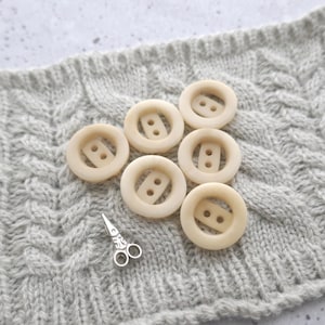 May include: Six off-white round buttons with two holes each, arranged in a semi-circle on a gray knitted fabric. A small pair of silver scissors is in the lower left corner of the image.
