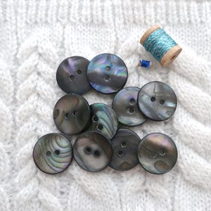 May include: A collection of iridescent, dark gray buttons with two holes each, arranged on a white cable-knit fabric. A wooden spool of teal thread and a small blue thread are also present, suggesting sewing or crafting.
