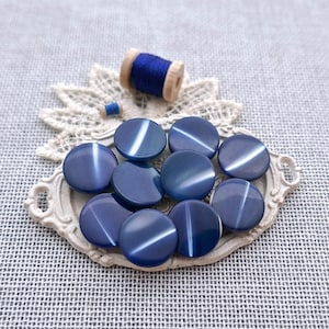May include: A collection of round, dark blue buttons with a smooth, reflective surface and a single light-colored horizontal stripe across the center of each. The buttons are arranged in a cluster on a decorative, off-white, oval-shaped tray.