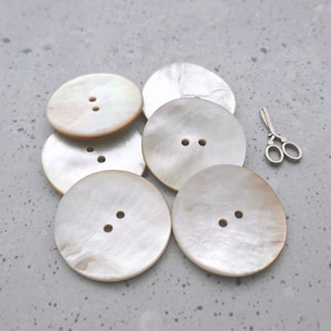 May include: A collection of large, round, pearlescent buttons with two holes each. The buttons are arranged on a gray surface, with a small pair of silver scissors nearby. The buttons have a subtle, iridescent sheen.
