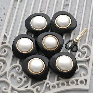 May include: Six black and white buttons with a gold trim and a pearl-like center. The buttons are irregularly shaped and arranged on a decorative white surface. A pair of gold and black scissors is also present.