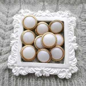 May include: A collection of white, round buttons with gold-colored rims, displayed in a white, ornate frame. The buttons have a glossy finish and are arranged on a gray, textured surface, suggesting a craft or sewing theme.