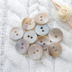 May include: A collection of round, iridescent buttons with two holes each. The buttons vary in color, with shades of white, beige, and pale blue. They are arranged on a white, textured surface, possibly fabric. The buttons are likely for sewing or crafting.