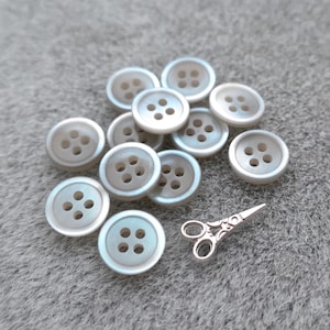 May include: A collection of iridescent, pearlescent buttons with four holes each, scattered on a gray surface. A small, silver-colored pair of decorative scissors is also present, suggesting a sewing or crafting theme.