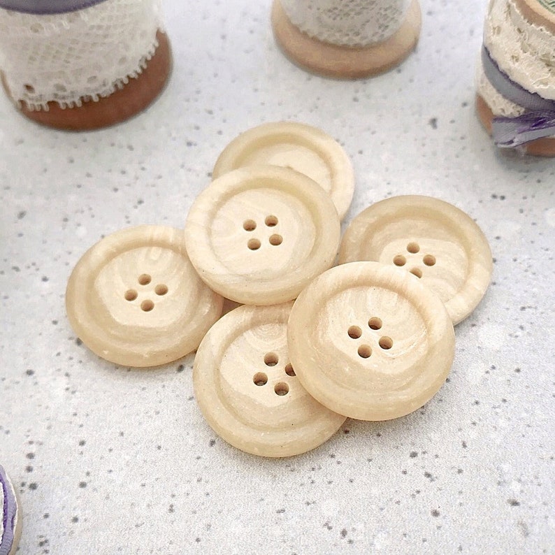 Puede incluir: Seis botones de madera beige claro, cada uno con cuatro agujeros. Los botones tienen una superficie texturizada y un dise&ntilde;o circular. Est&aacute;n dispuestos en un mont&oacute;n suelto, adecuados para coser o manualidades. Cada bot&oacute;n mide aproximadamente 2,5 cm de di&aacute;metro.