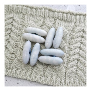 May include: Six light blue ceramic buttons with a white swirl design. The buttons are shaped like a long oval with a hole in the center. The buttons are arranged in a circle on a light gray knitted fabric.