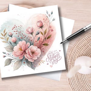 Puede incluir: Una tarjeta del Día de la Madre en acuarela con un diseño en forma de corazón en tonos rosas y azules. El corazón está adornado con flores rosas, hojas verdes y la frase "Happy Mother's Day". También se ve un bolígrafo y un objeto decorativo.