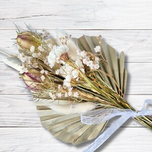 Puede incluir: Un ramo de flores secas en tonos blancos y beige, atado con una cinta blanca y arreglado en un abanico de hojas de palma secas.