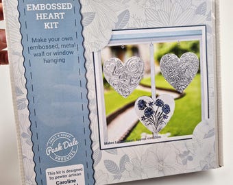May include: A craft kit box with the text "Embossed Heart Kit" and "Make your own embossed, metal wall or window hanging." The box features a picture of three heart-shaped decorations. The kit is designed by pewter artisan Caroline Buchwald.