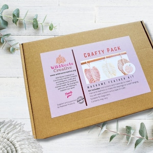 May include: A brown cardboard box with a pink and white label that says "Crafty Pack Macrame Feather Kit". The label includes instructions and materials to make 3 macrame feathers. The box is on a white wooden surface.