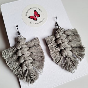May include: A pair of grey macrame earrings with a tassel fringe. The earrings are hanging on a white card with a butterfly logo that says "Shining Bright Arts, Crafts & Illustration".