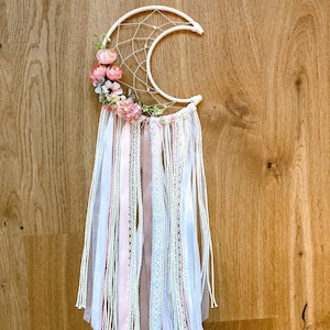 Könnte beinhalten: Ein weißer Halbmond-Traumfänger mit rosa und weißen Bändern und floralen Akzenten.