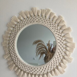 May include: A round mirror with a white macrame frame and gold beads. The frame is decorated with white tassels.