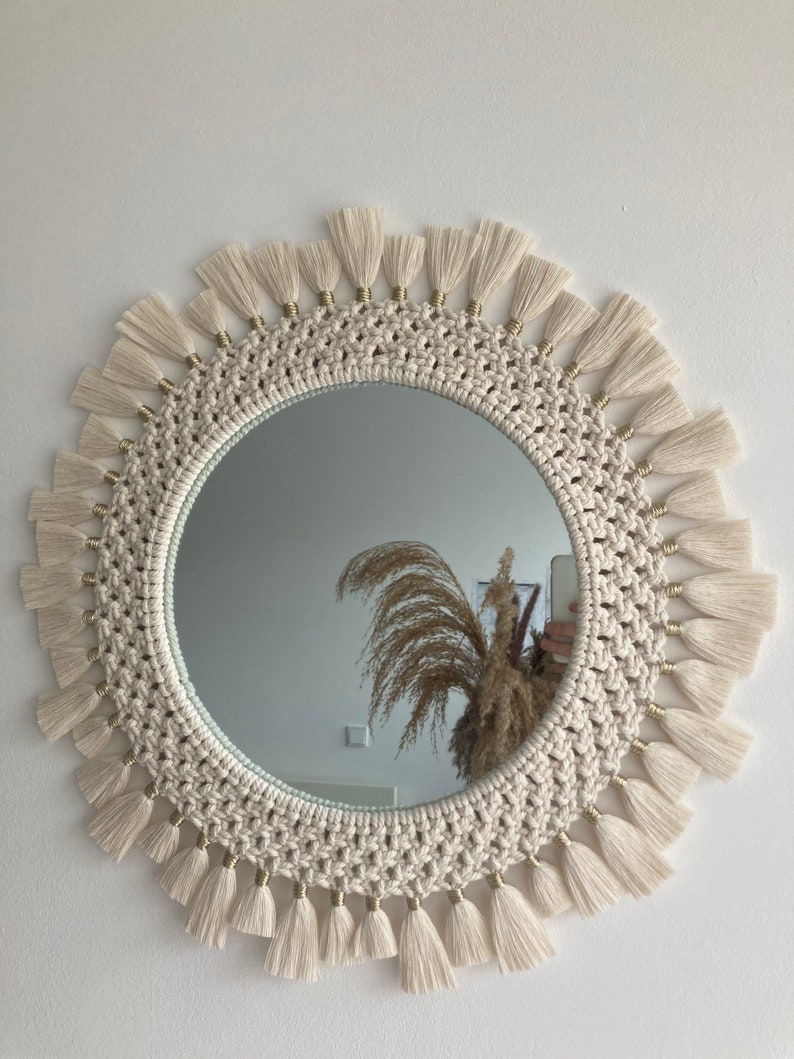 May include: A round mirror with a white macrame frame and gold beads. The frame is decorated with white tassels.