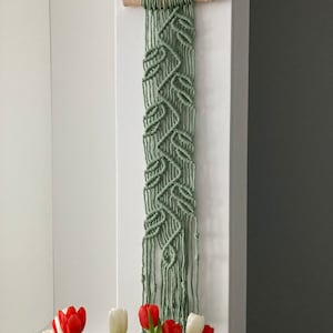 Puede incluir: Un tapiz de pared de macramé verde salvia con un patrón de hojas, suspendido de una clavija de madera natural. La obra de arte textil hecha a mano se exhibe contra una pared blanca, con un ramo de tulipanes rojos y blancos en primer plano.