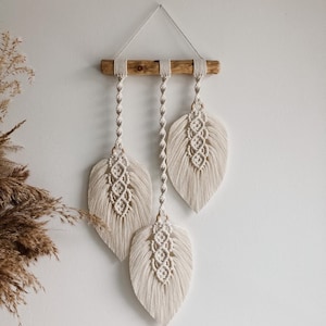 Può includere: Un arazzo da parete con tre forme di piume in macramé appese a un bastone di legno. Le piume sono di colore crema e hanno un dettaglio a frangia.