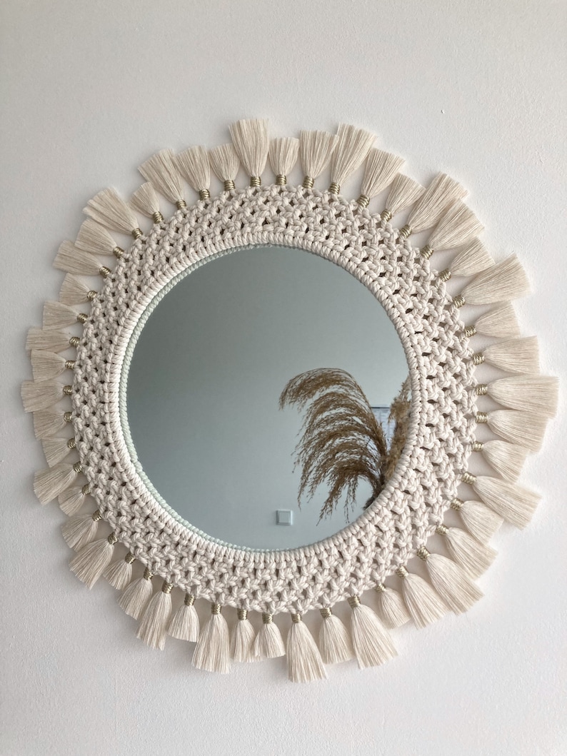 May include: A round mirror with a macrame frame and white tassels. The mirror has a gold trim around the edge.