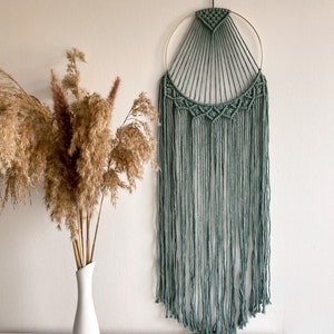 Könnte beinhalten: Ein Wandbehang aus Makramee in Salbeigrün mit einem goldenen Ring und langen Fransen. Der Behang befindet sich an einer weißen Wand mit einer Vase mit getrocknetem Pampasgras davor.