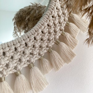 May include: A cream-colored macrame wall hanging with a crescent moon shape and gold accents. The hanging has a fringe of tassels along the bottom edge.