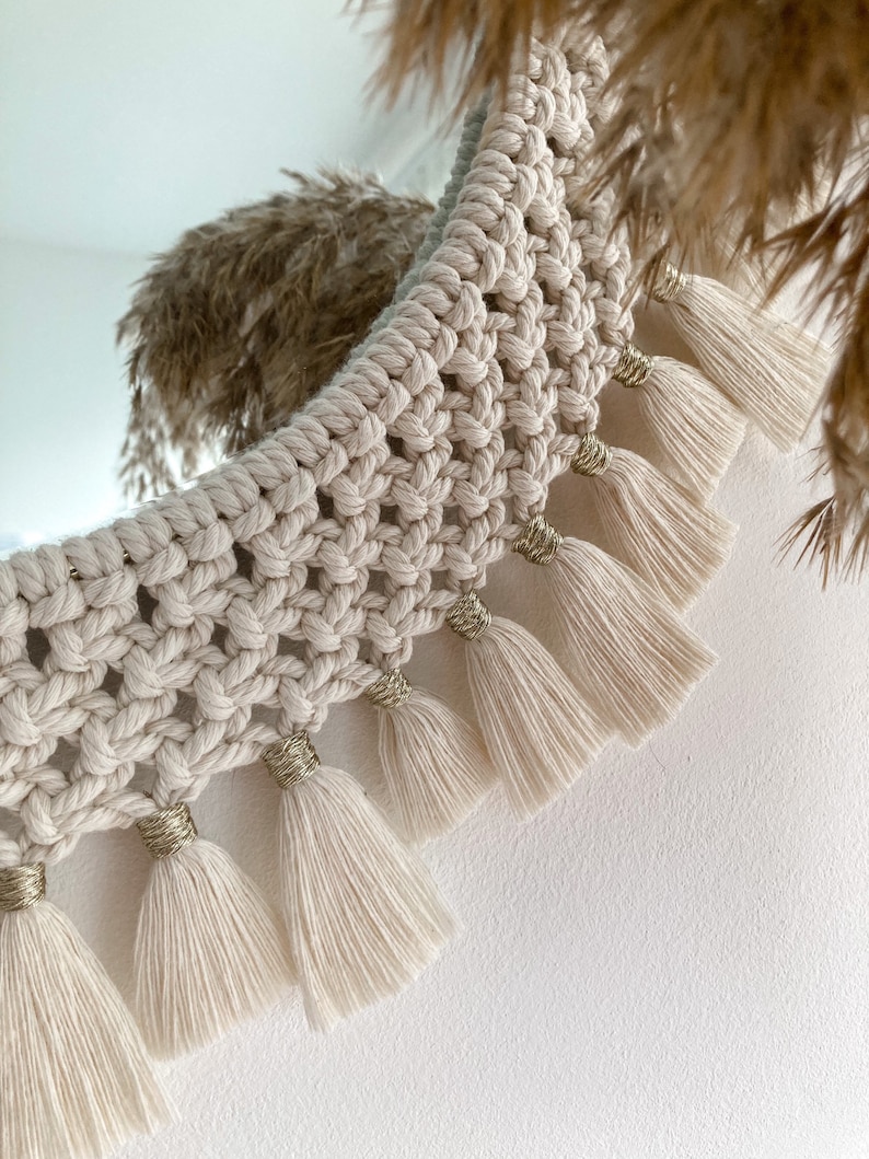 May include: A cream-colored macrame wall hanging with a crescent moon shape and gold accents. The hanging has a fringe of tassels along the bottom edge.