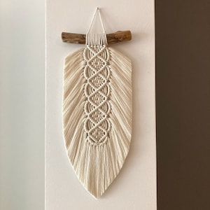 May include: Cream-colored macrame wall hanging with a central diamond pattern. The decorative piece is suspended from a natural wood branch. The macrame design features a textured, woven pattern and a fringed bottom, creating a bohemian aesthetic.