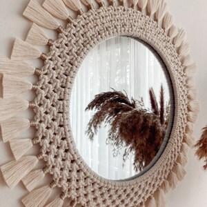 May include: A round mirror with a macrame frame and a fringe border. The frame is made of natural beige rope and the fringe is made of white yarn.