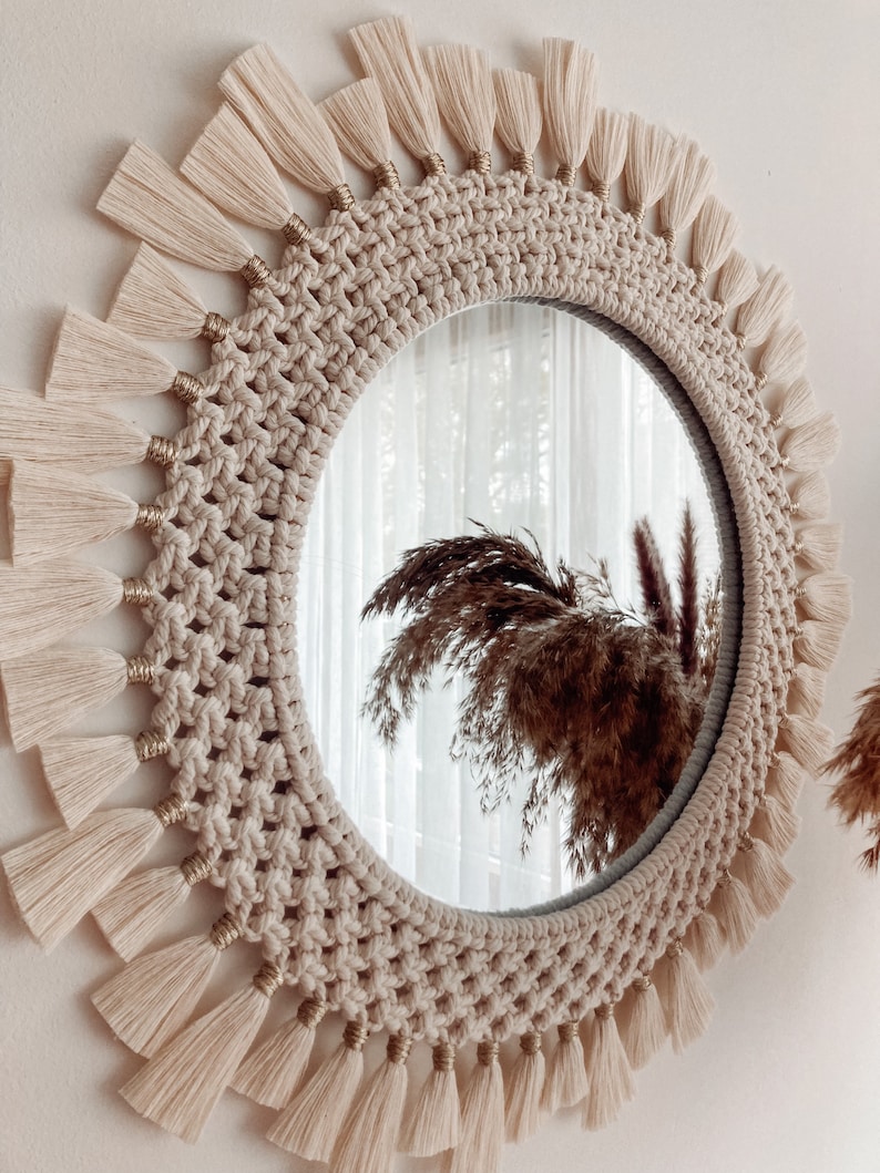 May include: A round mirror with a macrame frame and a fringe border. The frame is made of natural beige rope and the fringe is made of white yarn.