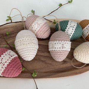 Könnte beinhalten: Sechs handgefertigte Ostereier, verziert mit weißer Spitze und Schnur. Die Eier sind in verschiedenen Farben, darunter rosa, beige, braun und grün.