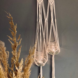 May include: Two white macrame plant hangers with glass candle holders. The candle holders are hanging from the macrame and have a white tassel at the bottom.