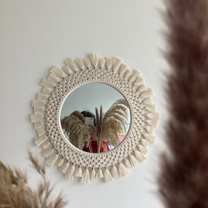 May include: A round mirror with a white macrame frame and tassels. The mirror is hanging on a white wall.